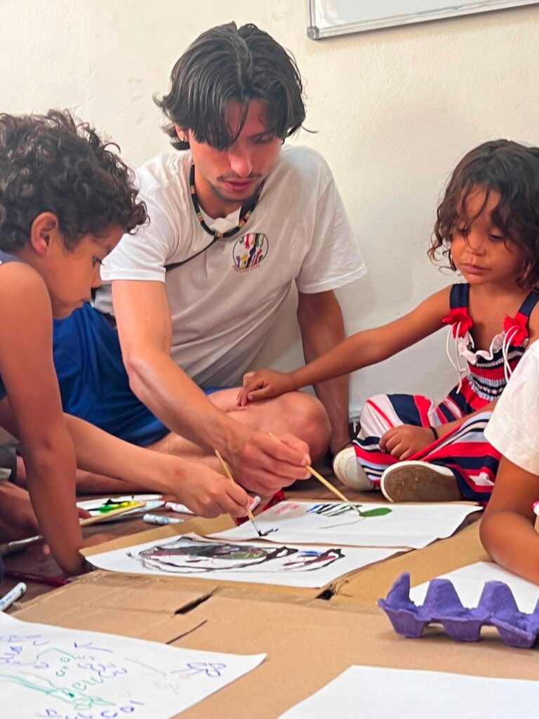 Un volontario dipinge su grandi fogli di carta insieme a due bambini seduti a terra