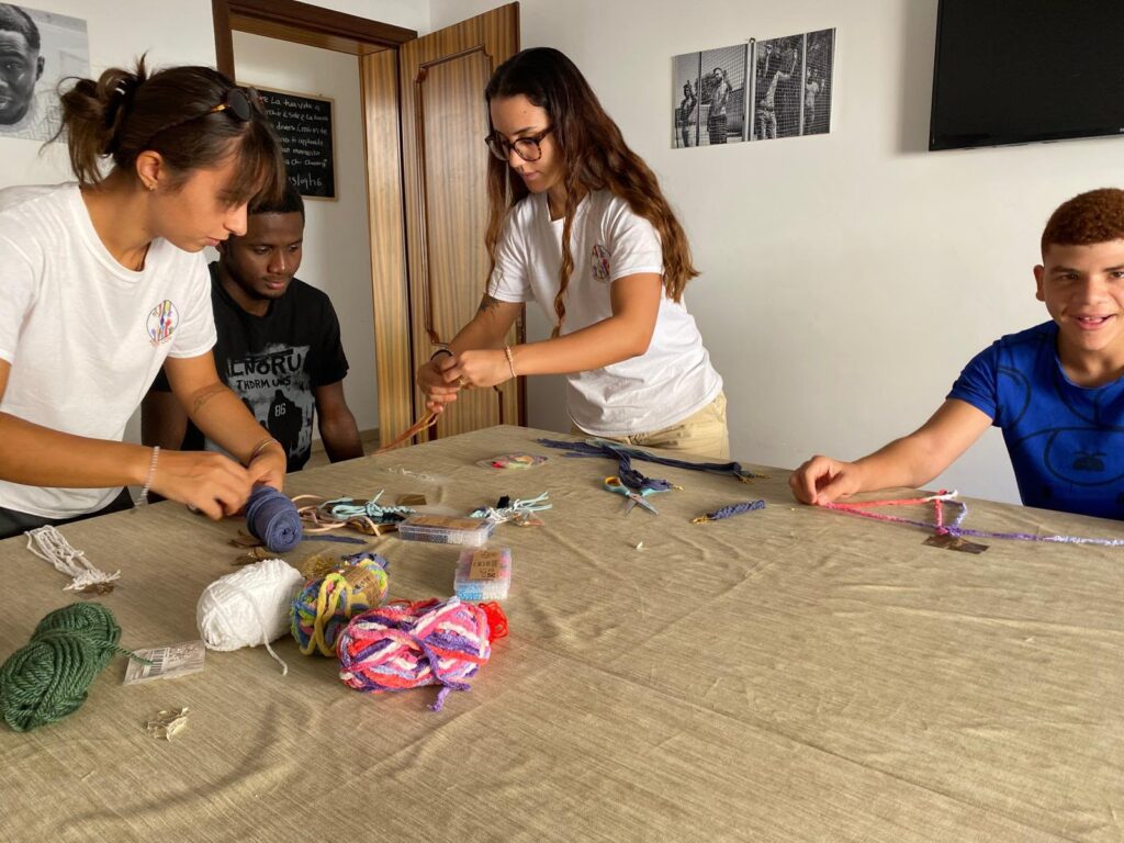 Volontarie e ragazzi intrecciano braccialetti di filo attorno a un tavolo